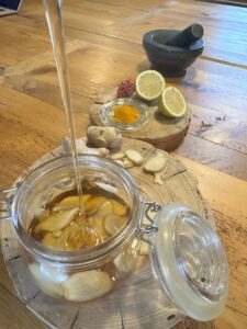 Auf einem rustikalen Holztisch steht ein geöffnetes Glas mit Ingwerscheiben in einer goldgelben Flüssigkeit. Aus einer schmalen Tülle fließt gerade ein dünner Strahl Honig in das Glas. Daneben liegen frische Ingwerwurzeln und einige Ingwerscheiben auf einer runden Holzscheibe. Im Hintergrund stehen eine halbierte Zitrusfrucht, eine kleine Glasschale mit leuchtend gelbem Kurkumapulver, rötliche Beeren und ein dunkler Steinmörser mit Stößel. Das natürliche Licht und die warmen Holztöne verleihen dem Bild eine gemütliche, handwerkliche Atmosphäre.