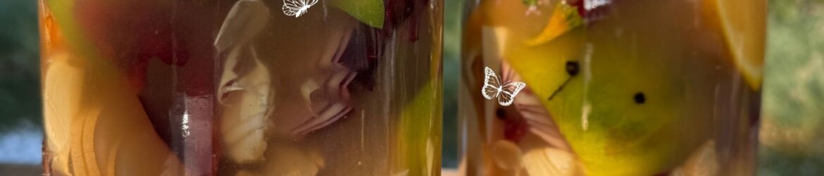 Zwei große Gläser mit selbst angesetztem Fire Cider stehen im Sonnenlicht. Im goldbraunen Apfelessig sind Stücke von Zitrusfrüchten, Zwiebel, Knoblauch, Ingwer, Kräutern und Gewürzen zu erkennen; darüber steht der Schriftzug „Firecider“ und das Logo der Wildkräuterwerkstatt.